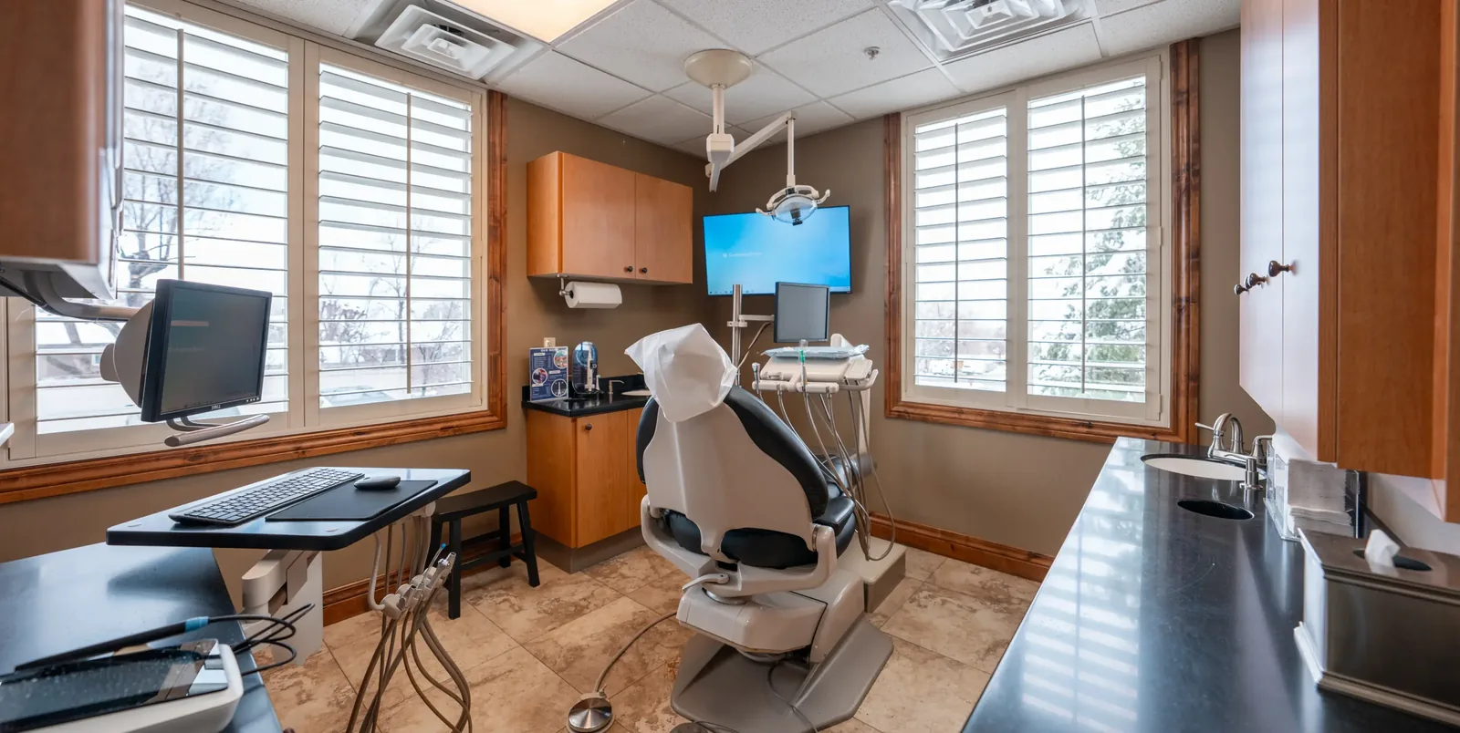 Dental operatory with an examination chair, medical equipment, monitors, and shuttered windows.