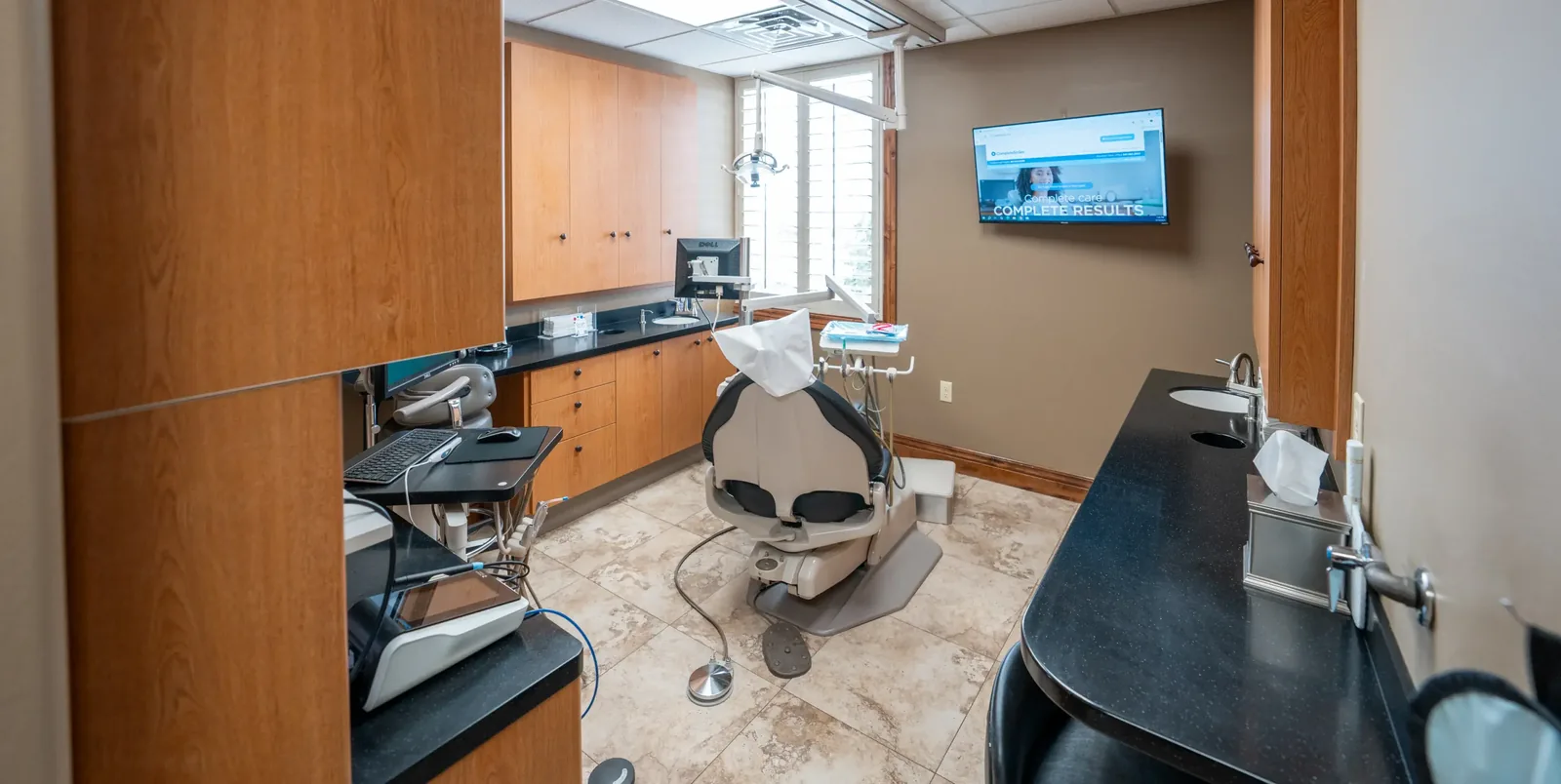 Dental operatory room with an examination chair, wooden cabinetry, and a wall-mounted television.